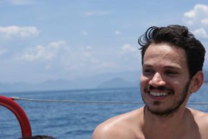 A smiling person named Emmanuel on a boat with the blue ocean and distant mountains under a clear sky, expressing pure joy.