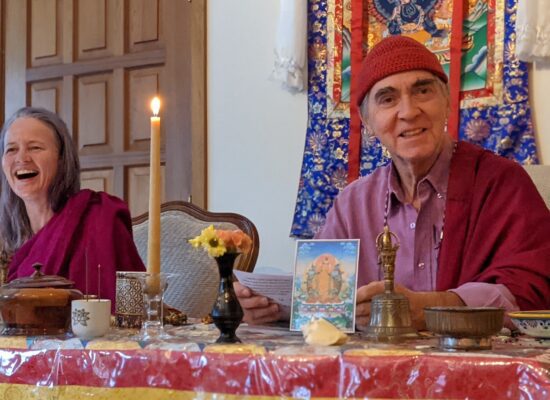 Two people in vibrant traditional robes smile at a table with a candle and flowers in a joyful setting.
